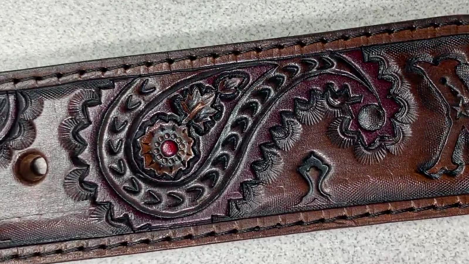 A close-up of a brown tooled leather belt featuring an embossed paisley pattern with a small red gemstone accent in the center, resting on a light gray textured surface.